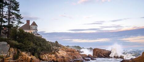 A coastal house with a prominent turret, situated on a rocky shoreline with waves crashing against the rocks.