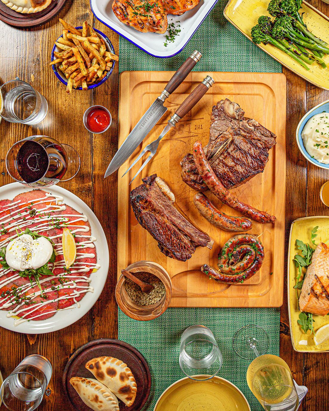 Una mesa de madera con varios platos, incluyendo filete, salchichas y una ensalada.