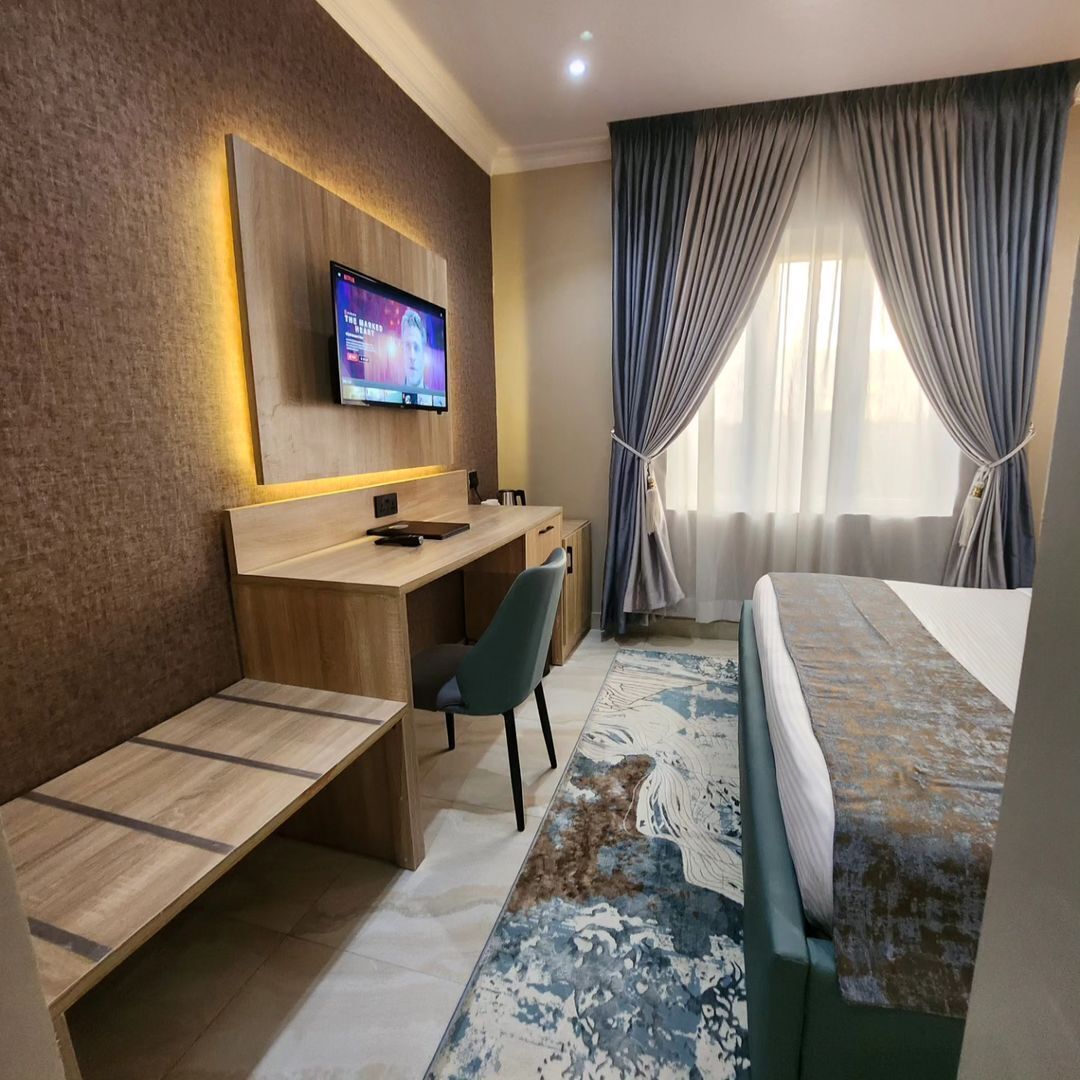 A hotel room with a bed, a desk, a chair, a television mounted on the wall, and a window with curtains.