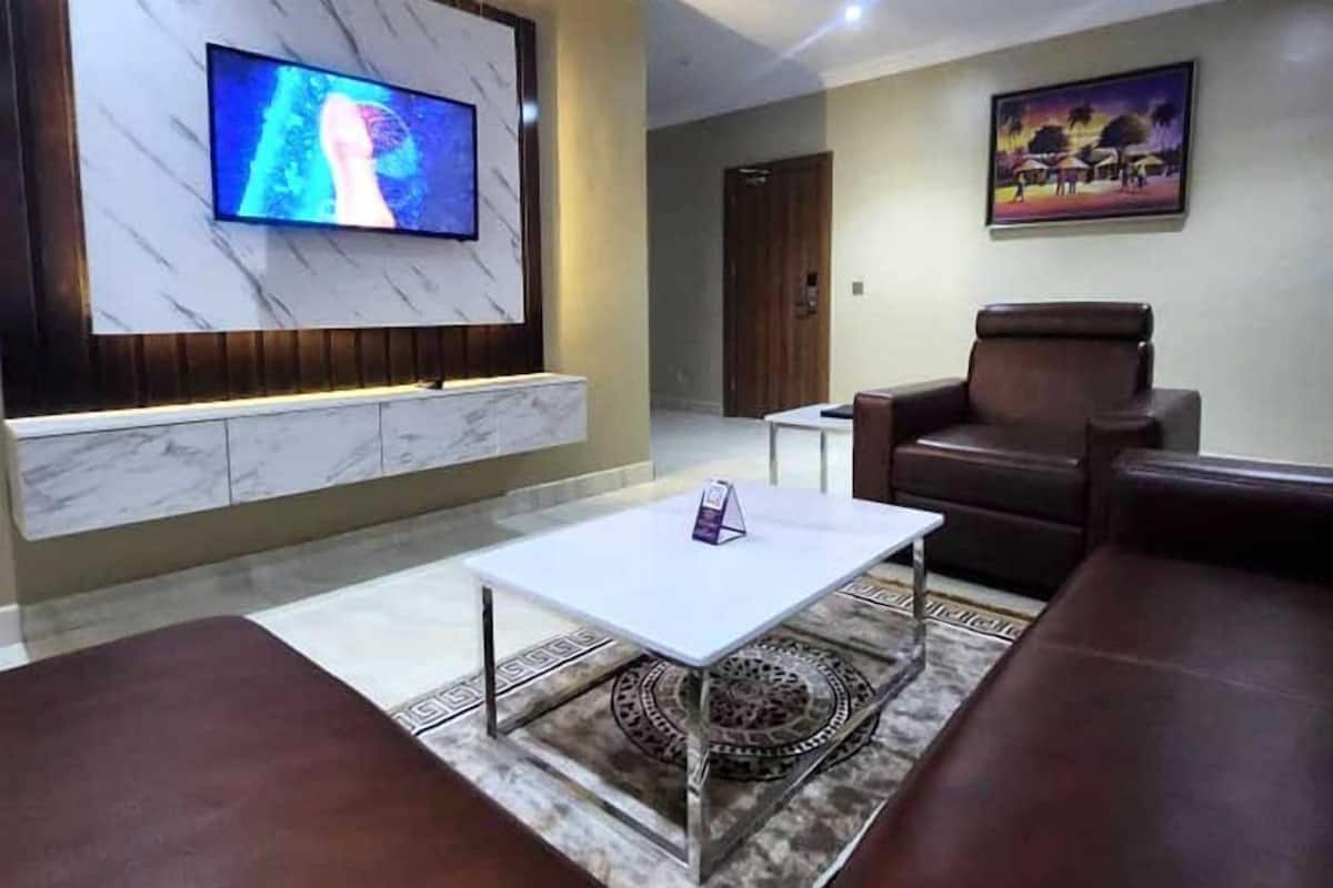 A modern living room with a flat-screen TV mounted on a marble wall, a white coffee table, a brown leather sofa, and a patterned rug.