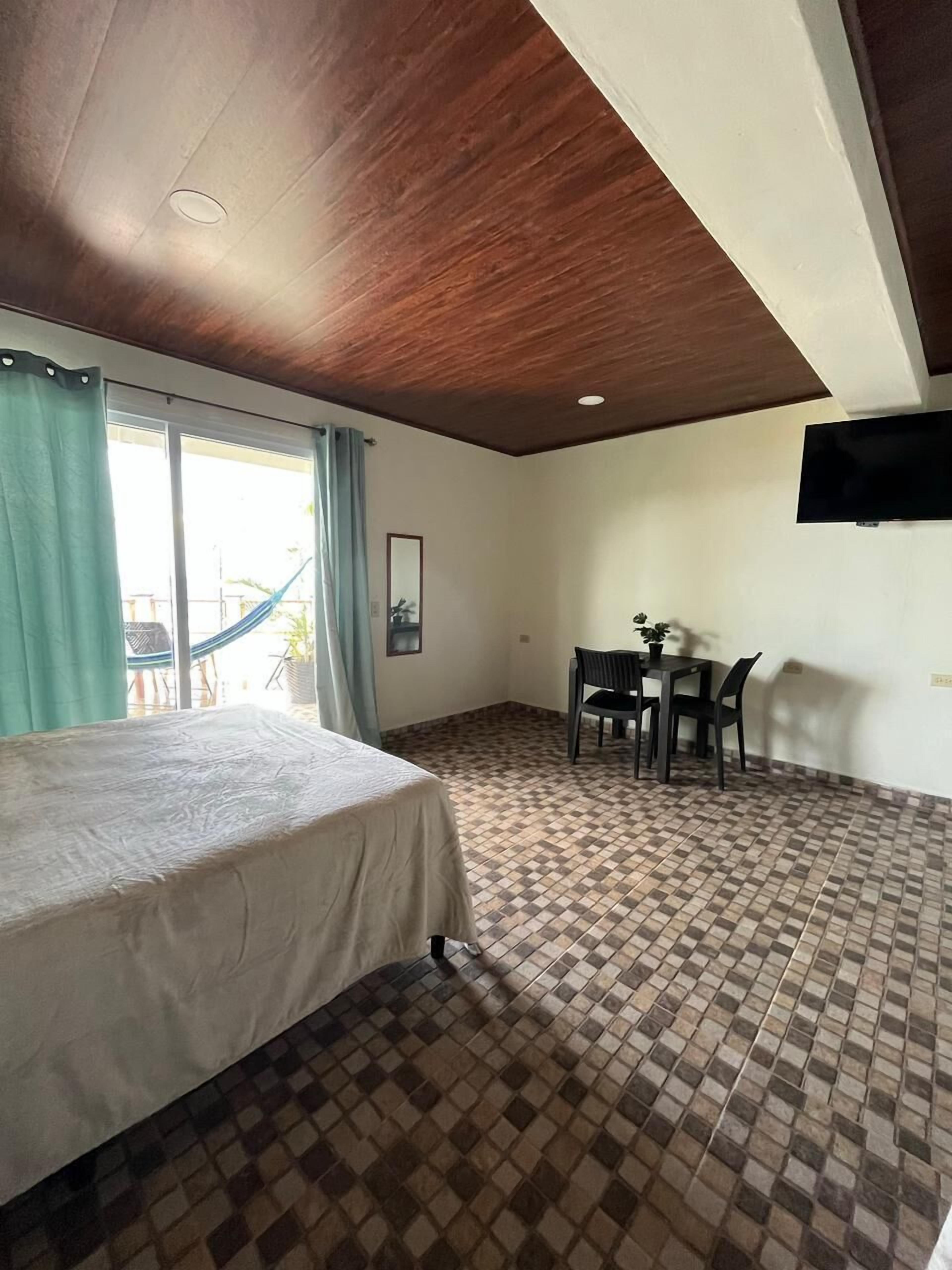 A hotel room with a bed, a dining table with chairs, a TV mounted on the wall, and a view through a window.