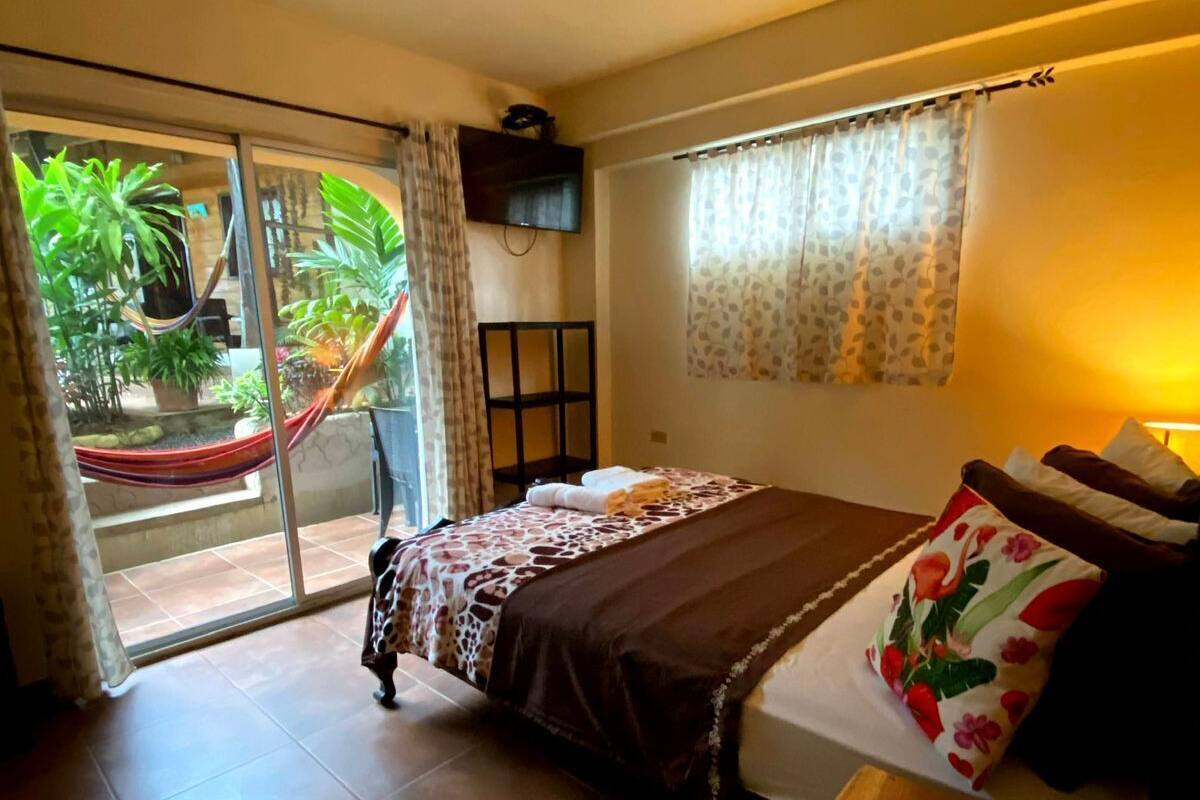A bedroom with a bed, a TV, a window with curtains, a small table with a remote, and a view of a garden through a glass door.
