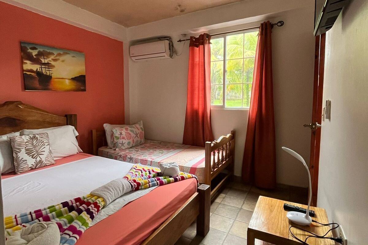A bedroom with two beds, a desk, a chair, a window with curtains, and an air conditioning unit.