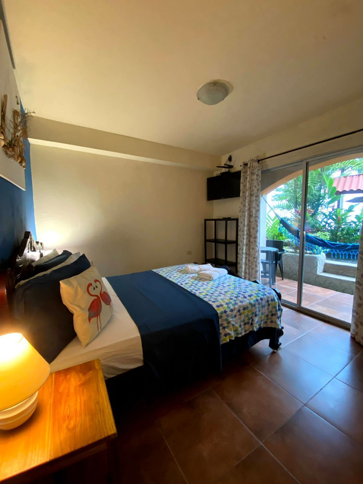 Una habitación de hotel con cama, un escritorio con lámpara, televisión y puerta corrediza de vidrio a un área exterior con plantas.