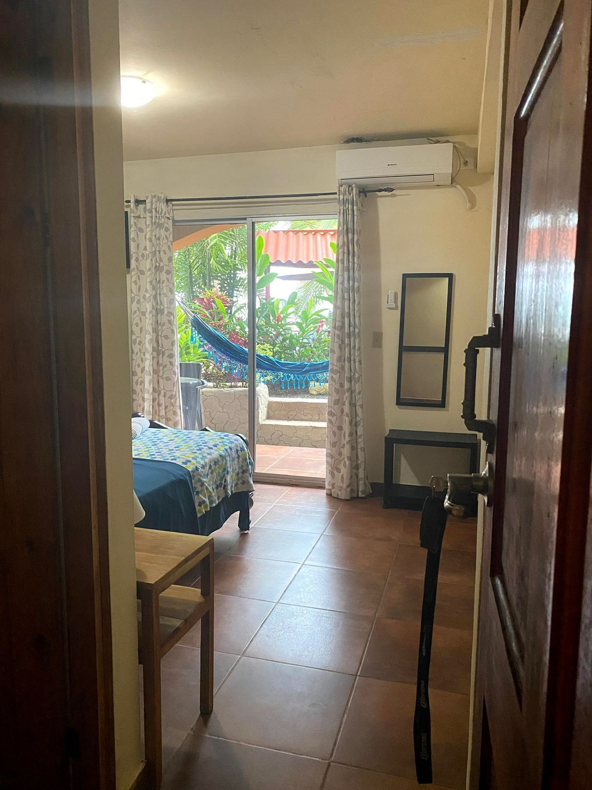 A hotel room with a bed, a small table, a chair, a sliding glass door leading to a balcony with a hammock, and an air conditioning unit on the wall.
