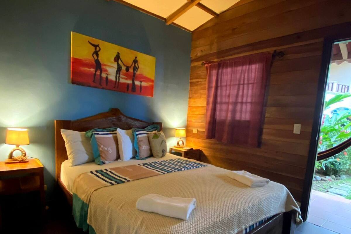 A bedroom with a wooden ceiling, a bed with multiple pillows, a painting on the wall, and a window with curtains.