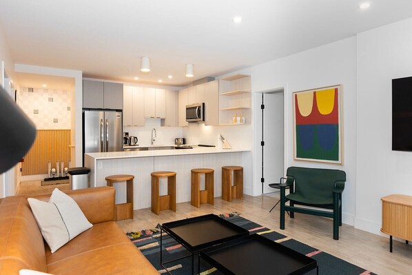A modern kitchen with white cabinets, a stainless steel refrigerator, and a microwave. There are wooden stools and a green chair in the living area.
