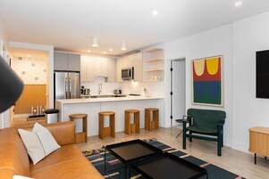Una cocina moderna con armarios blancos, un refrigerador de acero inoxidable y un microondas. Hay taburetes de madera y una silla verde en la sala de estar.