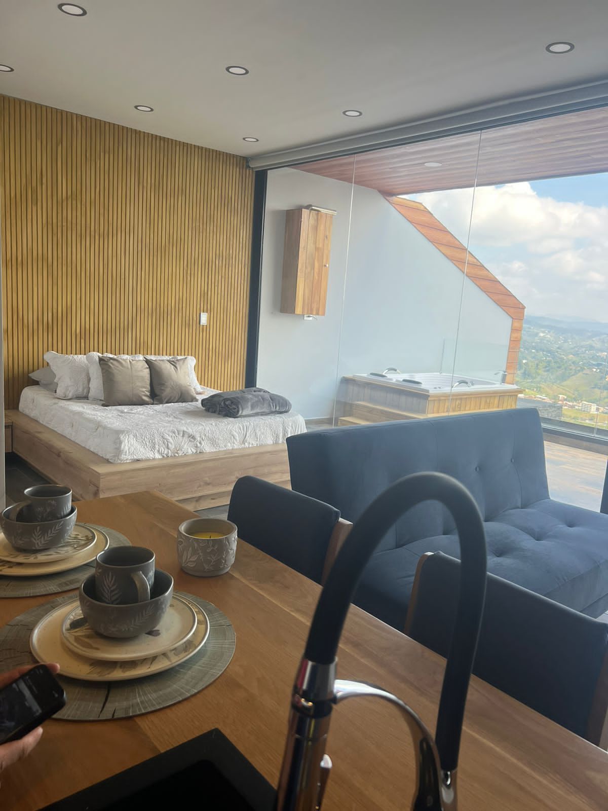 Un dormitorio moderno con una cama grande, una zona de comedor con mesa y sillas, y vistas al paisaje a través de un ventanal.