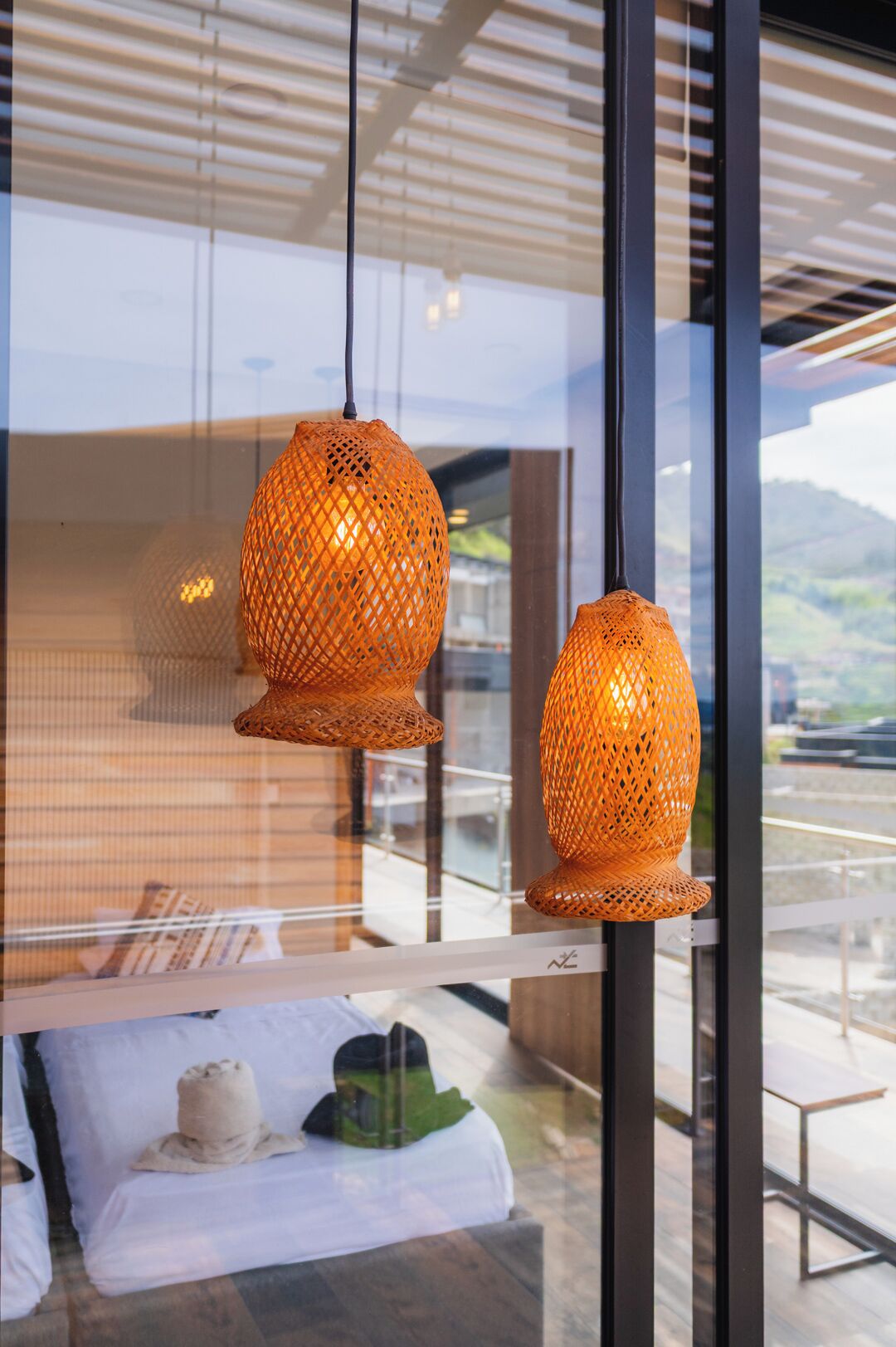 A modern interior with two spherical wicker lampshades hanging from the ceiling, a glass table with a white cloth and a green hat, and a view of a mountainous landscape through large windows.