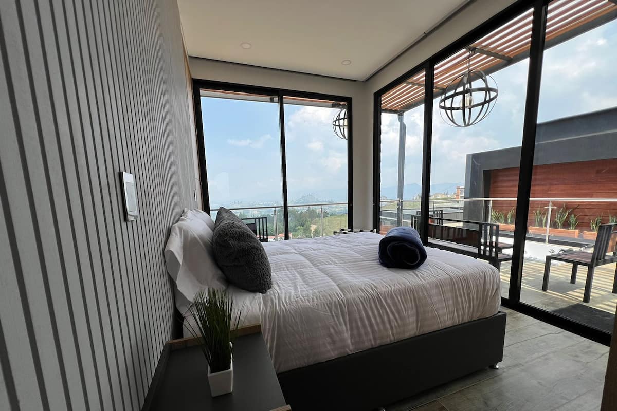 Une chambre moderne avec un grand lit, une plante en pot et une vue sur un balcon aménagé avec du mobilier d’extérieur.
