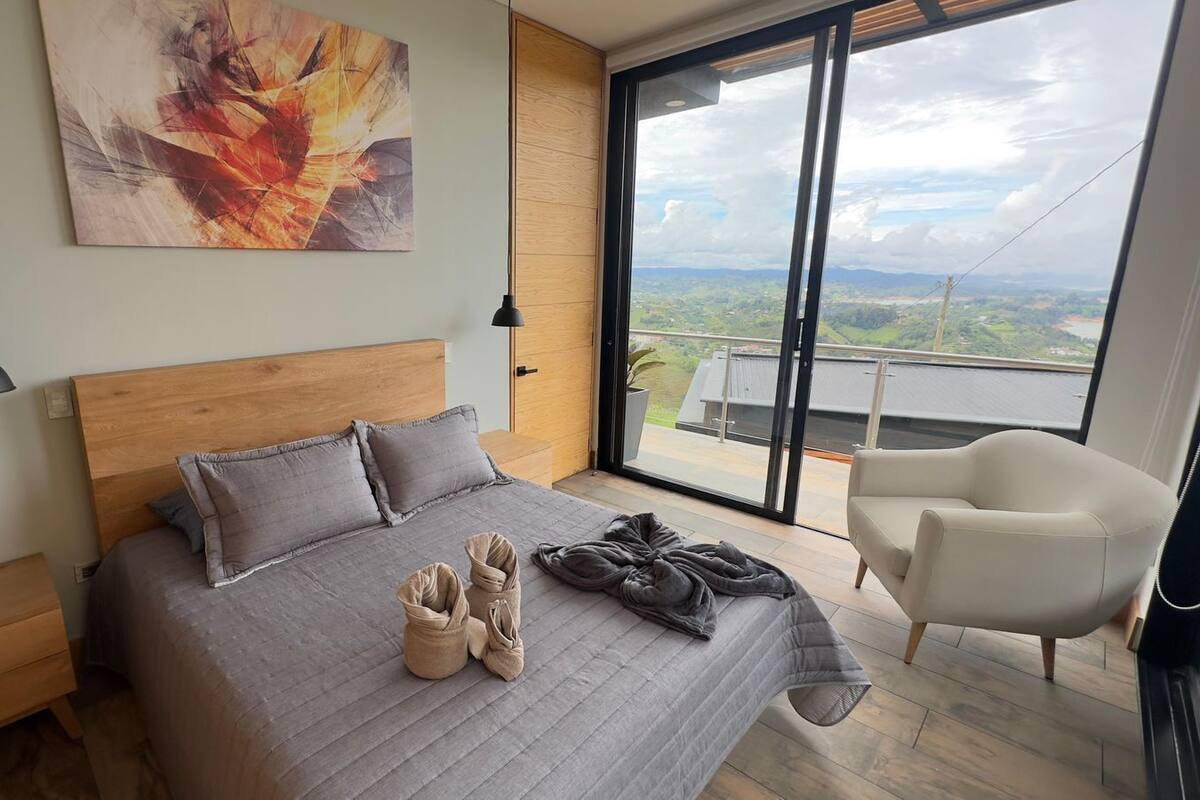 Une chambre avec un grand lit, une vue sur la campagne par une porte-fenêtre coulissante, un fauteuil moderne et un tableau au mur.