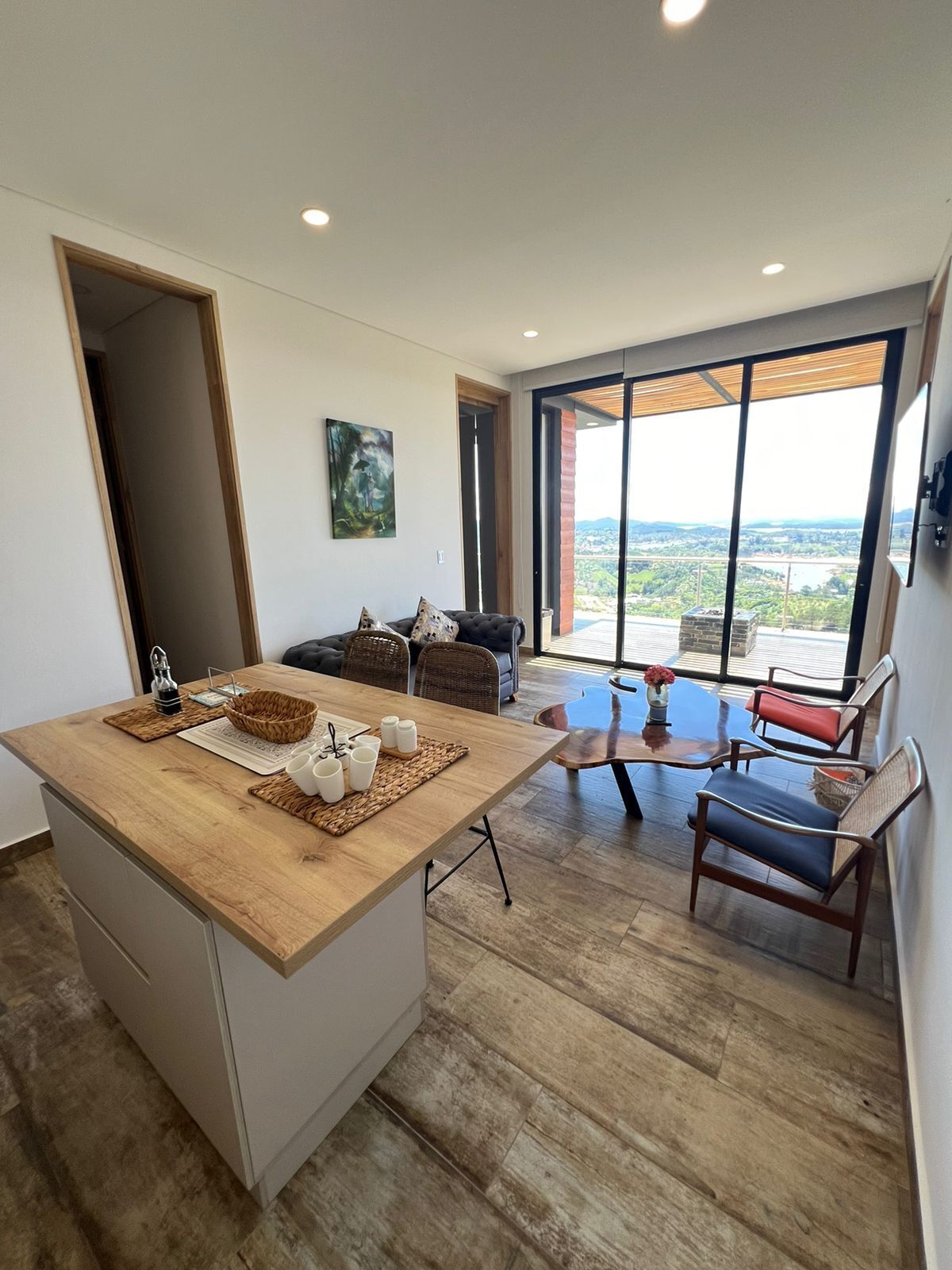 Una cocina moderna con una isla de madera, una zona de comedor con mesa de vidrio y vistas a un paisaje a través de ventanas amplias.