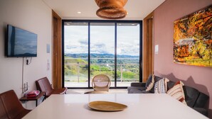 A modern room with a large dining table, a TV mounted on the wall, and a view of a landscape through large windows. - MONTECIELO HOSTING GUATAPE (El Peñol)