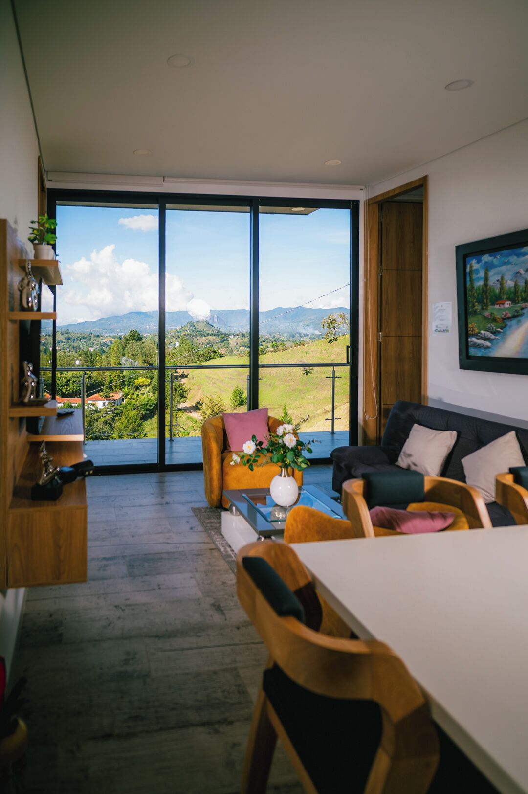Een moderne woonkamer met een grote schuifpui naar een balkon, een eethoek met houten meubels, en een schilderij aan de muur.