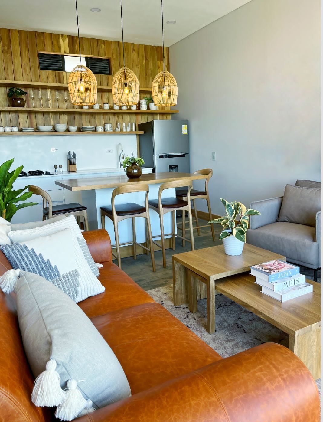 Una sala moderna con un sofá de cuero marrón, una mesa de centro de madera y un sofá gris. Hay lámparas colgantes de mimbre, una cocina al fondo y una planta en maceta sobre la mesa de centro.