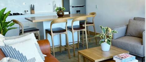 Una sala moderna con un sofá de cuero marrón, una mesa de centro de madera y un sofá gris. Hay lámparas colgantes de mimbre, una cocina al fondo y una planta en maceta sobre la mesa de centro.