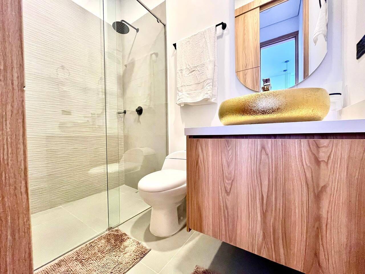 Un baño con cabina de ducha de vidrio, un inodoro blanco y un tocador de madera con lavamanos dorado.