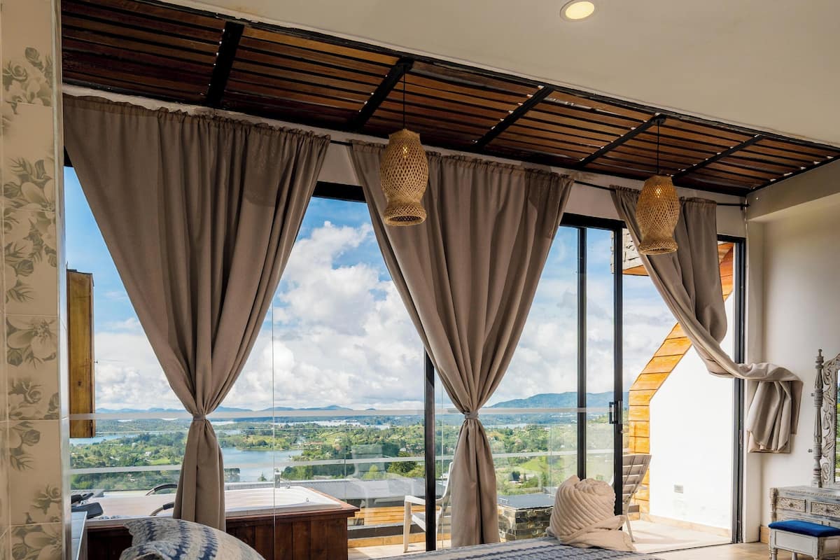 Une chambre avec un grand lit, deux fenêtres avec des rideaux, une vue sur un paysage et un plafond en bois.