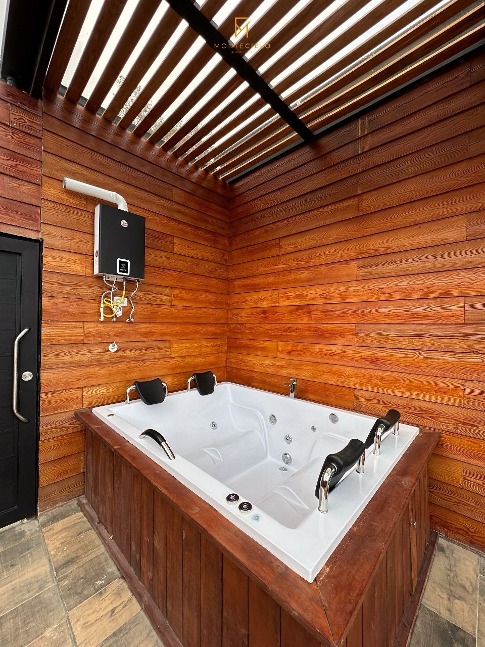 A wooden-paneled room with a large rectangular hot tub, a black door, and a ceiling with horizontal wooden slats.