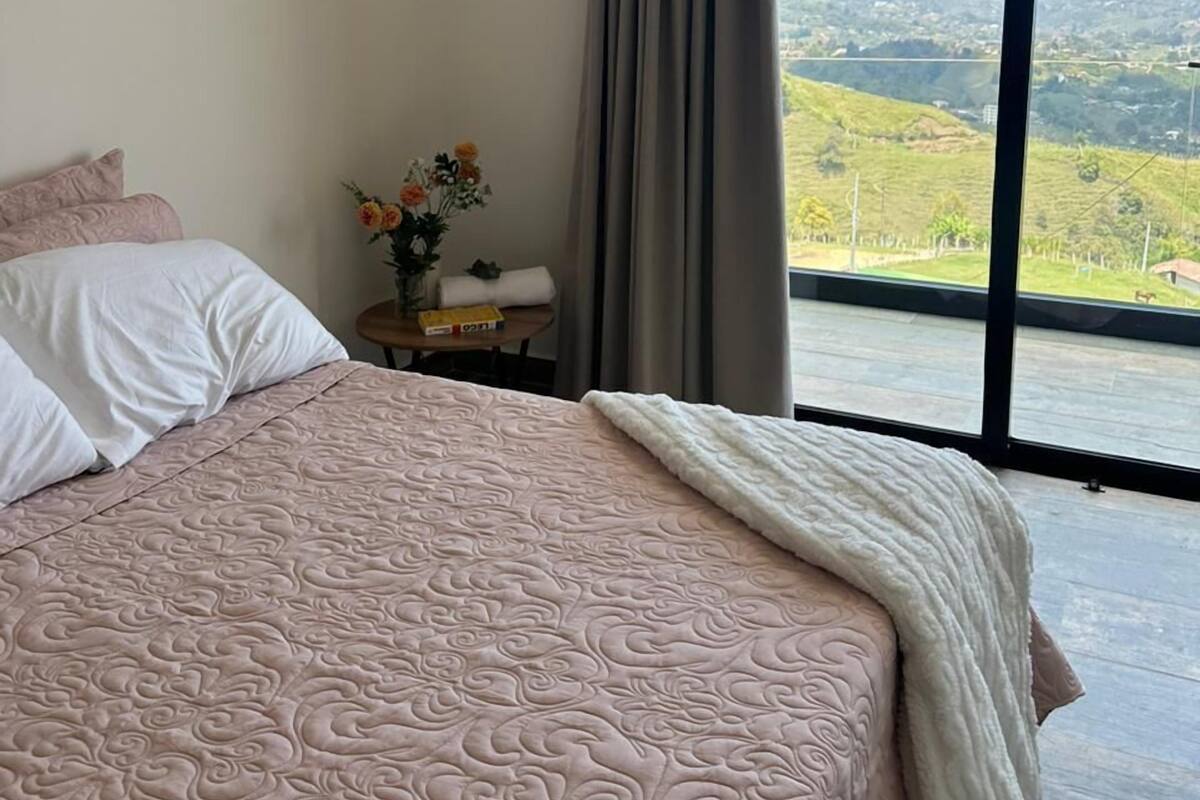 Une chambre avec un grand lit, une table de chevet ornée d’un vase de fleurs et une vue sur un paysage verdoyant, visible à travers une grande fenêtre.