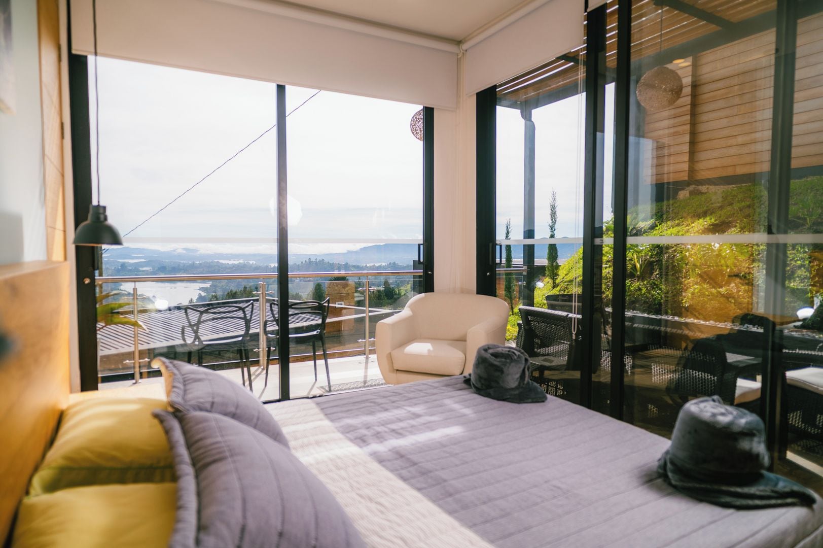 A balcony with a view of a body of water and hills, a bed with pillows, a chair, and a table.