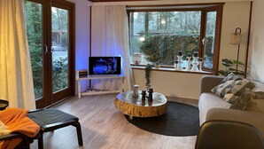 Una sala de estar con televisor sobre un mueble bajo, una mesa de centro redonda de madera, un sofá y un ventanal con vistas a árboles.