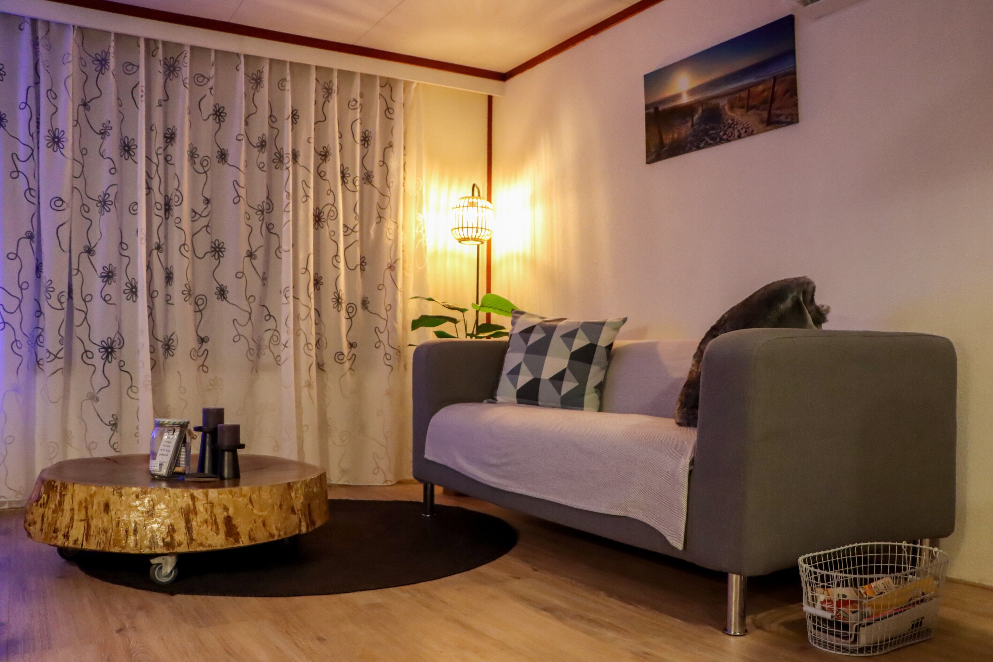 Un salón con un sofá gris, una mesa redonda de madera, una lámpara de pie y un cuadro en la pared.