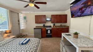 A compact hotel room with a bed, a ceiling fan, a kitchenette, a dining area, and a TV mounted on the wall.