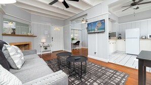 Un salon avec un canapé gris, une table basse noire et un tapis à motifs. On y trouve également un coin cuisine avec des meubles blancs et un réfrigérateur blanc.