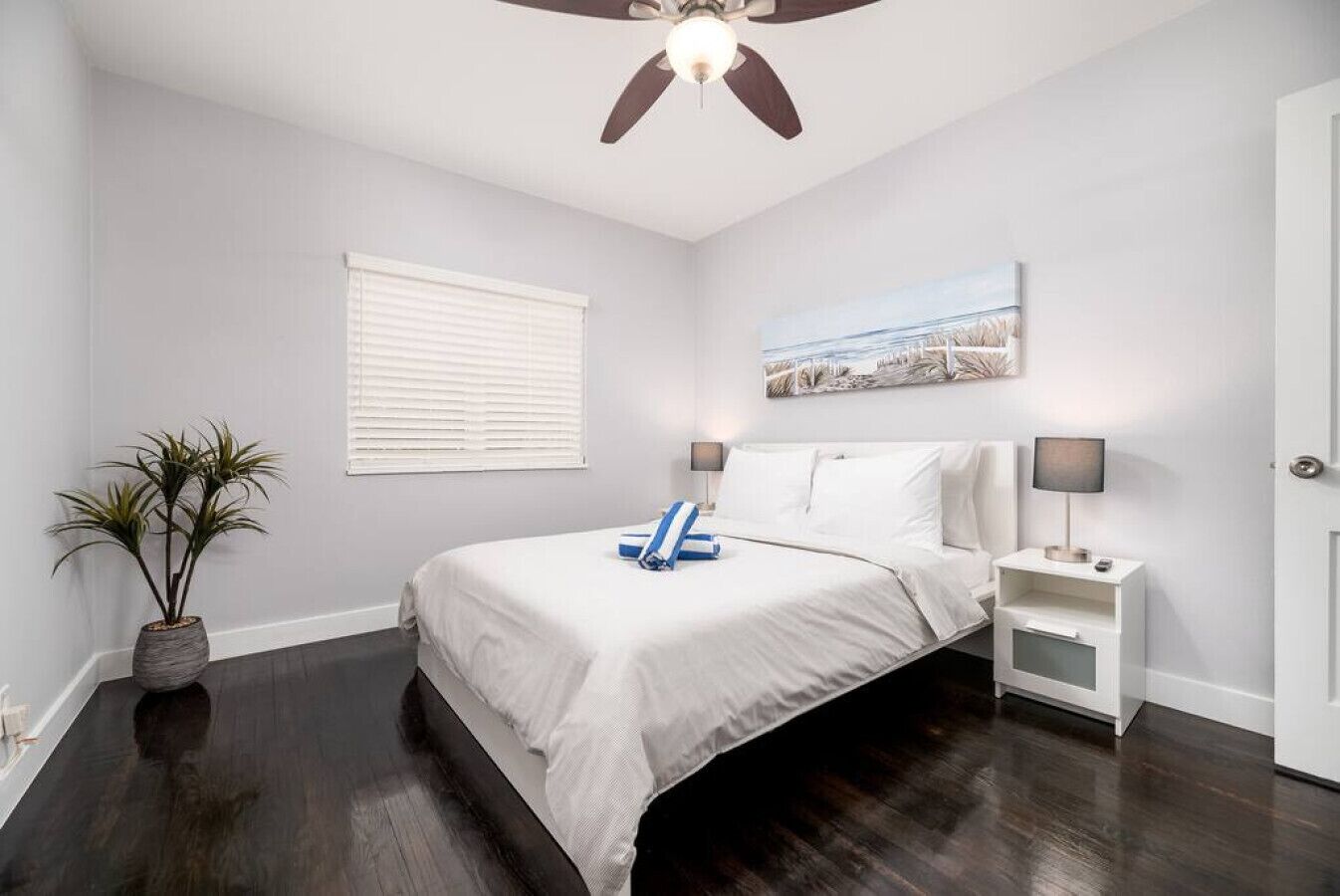 Quarto com cama, mesas de cabeceira, ventoinha no teto, janela com persianas, planta em vaso e um quadro na parede.