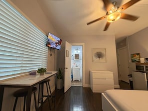A compact hotel room with a TV, ceiling fan, kitchenette, and a small bathroom.