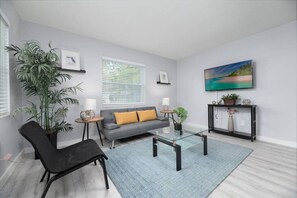 A modern living room with a grey sofa, a black chair, a glass coffee table, and a large window with blinds.