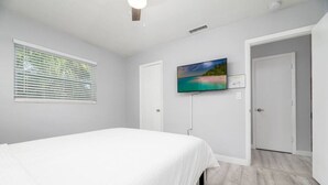 Um quarto com uma cama, uma televisão fixada na parede, uma janela com persianas e uma porta que dá para outro cômodo.