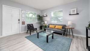 Uma sala de estar moderna com um sofá, uma mesa de centro de vidro, uma poltrona, uma mesinha lateral e uma porta.
