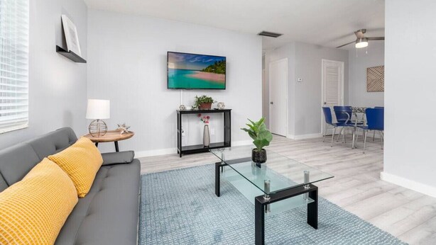Uma sala de estar moderna com um sofá cinza, uma mesa de centro de vidro, um tapete azul, um quadro na parede e uma área de jantar com cadeiras azuis.