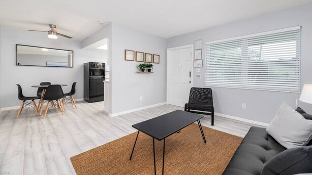 A modern living room with a black sofa, a dining area with a table and chairs, a black chair, a ceiling fan, and a window with blinds. - Short Walk to Wilton Drive! Apt2 (Wilton Manors)