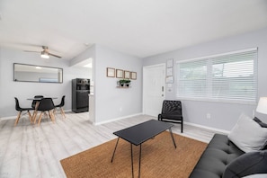 A modern living room with a black sofa, a dining area with a table and chairs, a black chair, a ceiling fan, and a window with blinds. - Short Walk to Wilton Drive! Apt2 (Wilton Manors)