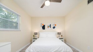 A bedroom with a bed, bedside tables, wall art, a ceiling fan, and a window with blinds.