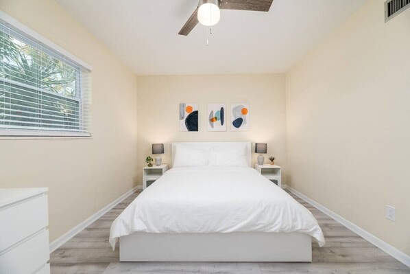 A bedroom with a bed, bedside tables, wall art, a ceiling fan, and a window with blinds.