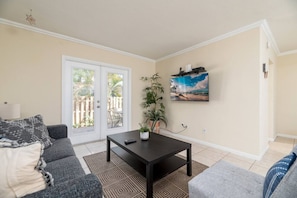 A living room with a sofa, a coffee table, a potted plant, a painting, and a sliding glass door leading to an outdoor area.