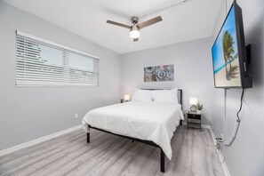 Ein Schlafzimmer mit einem Bett, einem Deckenventilator, einem an der Wand montierten Fernseher und einem Fenster mit Jalousien.
