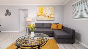 A living room with a grey sofa, a round glass coffee table, a vase with yellow flowers, and a large abstract painting on the wall. - Short Walk to Wilton Drive! Apt4 (Wilton Manors)