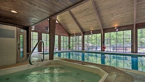 A large indoor swimming pool with a wooden interior, surrounded by large windows offering views of the outdoors.