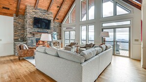 A spacious living room with a stone fireplace, large windows, and a high ceiling with wooden beams.