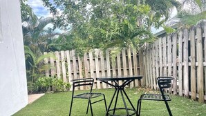Un pequeño patio al aire libre con una mesa y dos sillas, rodeado por un cerramiento de madera y follaje.