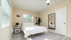 Un dormitorio con cama, mesitas de noche, un espejo, una ventana con persianas y una puerta.