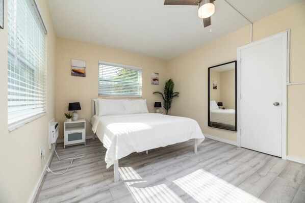 A bedroom with a bed, bedside tables, a mirror, a window with blinds, and a door.