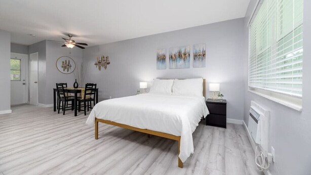 Une chambre à coucher comprenant un lit, un bureau, des chaises, un ventilateur de plafond, des œuvres d’art accrochées au mur et une fenêtre avec des stores.