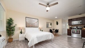 Une chambre comprenant un lit, des tables de chevet, un bureau, une chaise, une plante en pot, un ventilateur de plafond et une kitchenette équipée d’un four à micro-ondes et d’un four traditionnel.